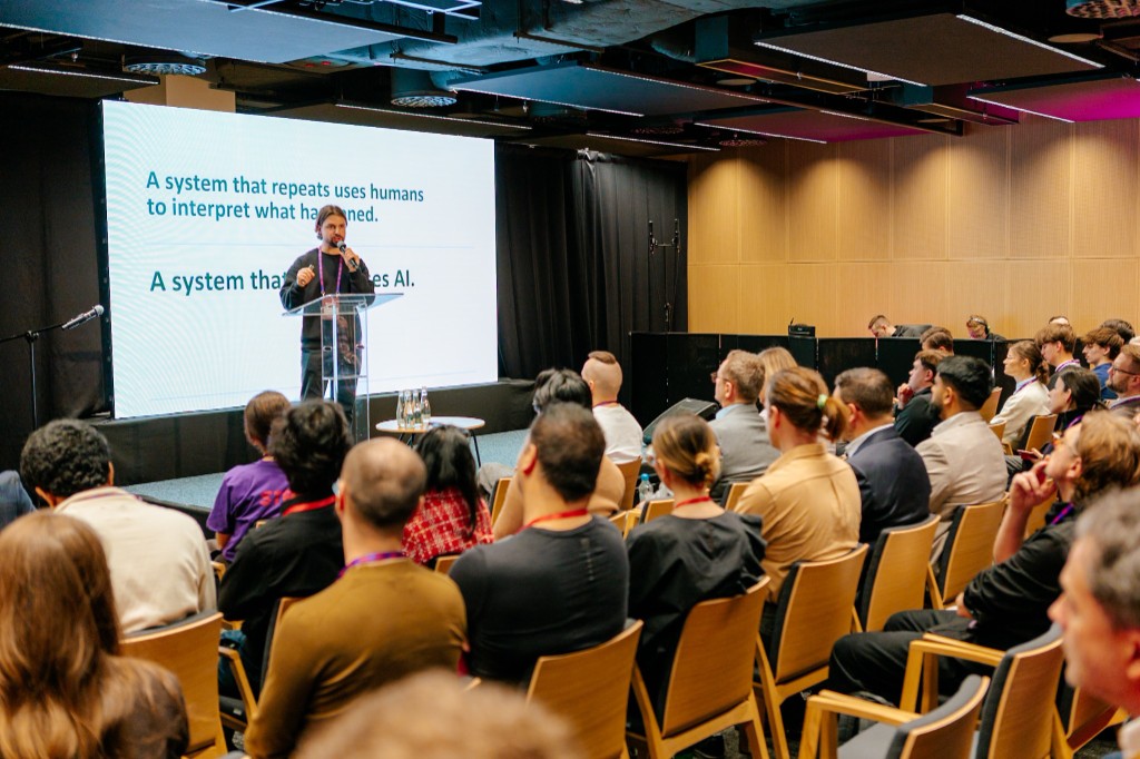 Macklin Buckler speaking from a lectern at MIT Universal AI in Warsaw. The slide behind him reads: a system that repeats uses humans to interpret what happened, a system that learns uses AI.